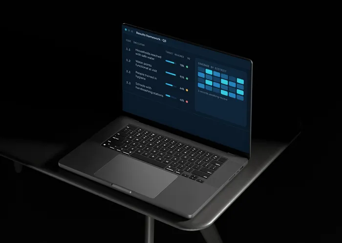 A laptop in a dark room showing an M&E results dashboard with sample data.